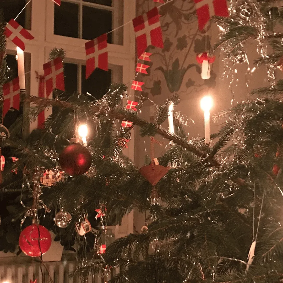Indoor Christmas tree with red baubles, tinsel, Danish flags, and lit candles.