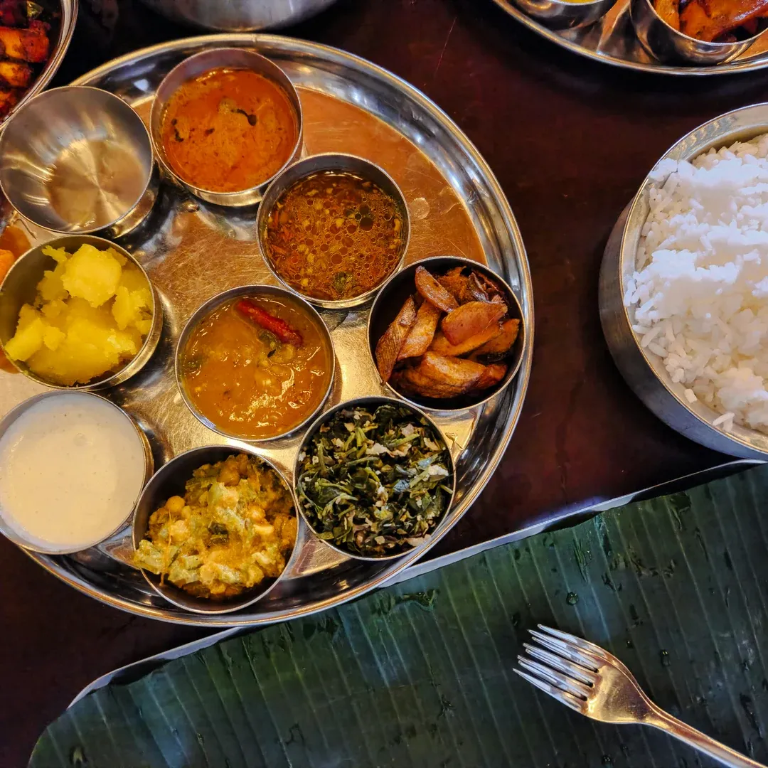 A metal thali with multiple small bowls of Indian curries and vegetables next to a bowl of white rice on a banana leaf placemat edge.