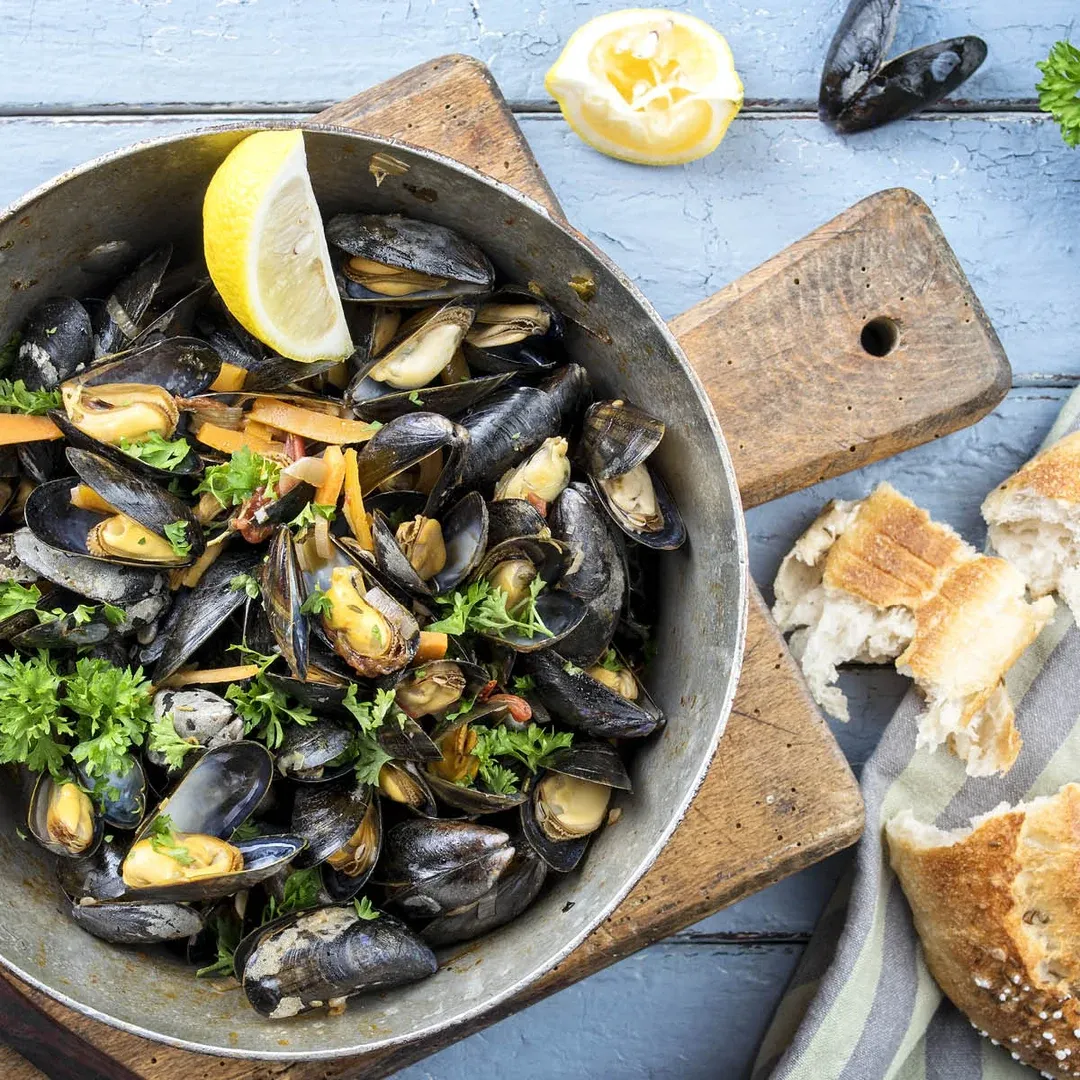 Mussels in a pan with parsley, lemon wedge, and crusty bread on a blue wooden table.