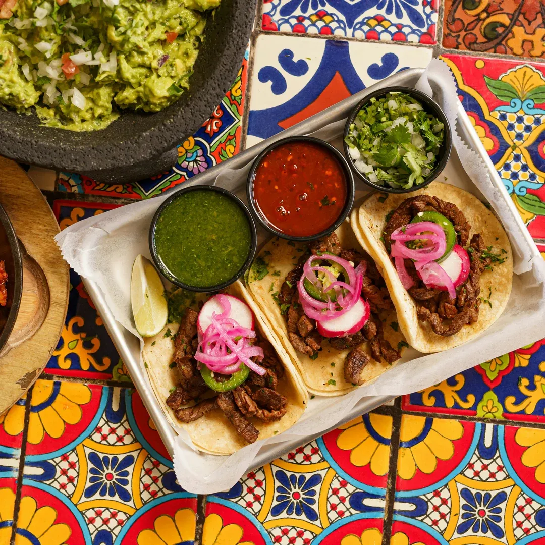 Three steak tacos topped with pickled onions, radish slices, and cilantro on warm tortillas, with green and red salsas nearby and guacamole on the side.