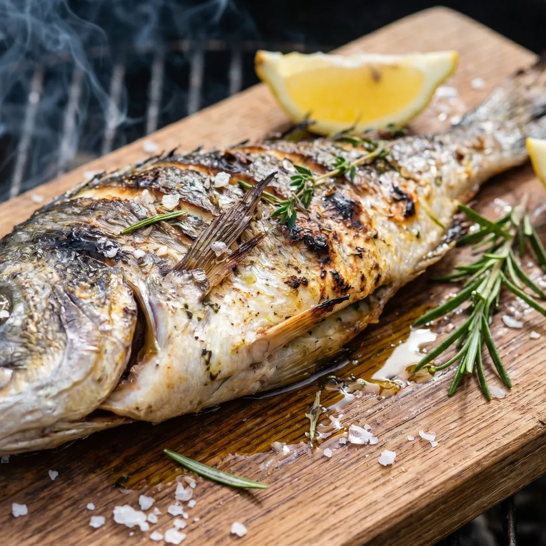 Grilled whole fish on a wooden board garnished with rosemary, thyme, and coarse salt, with lemon wedges in the background.