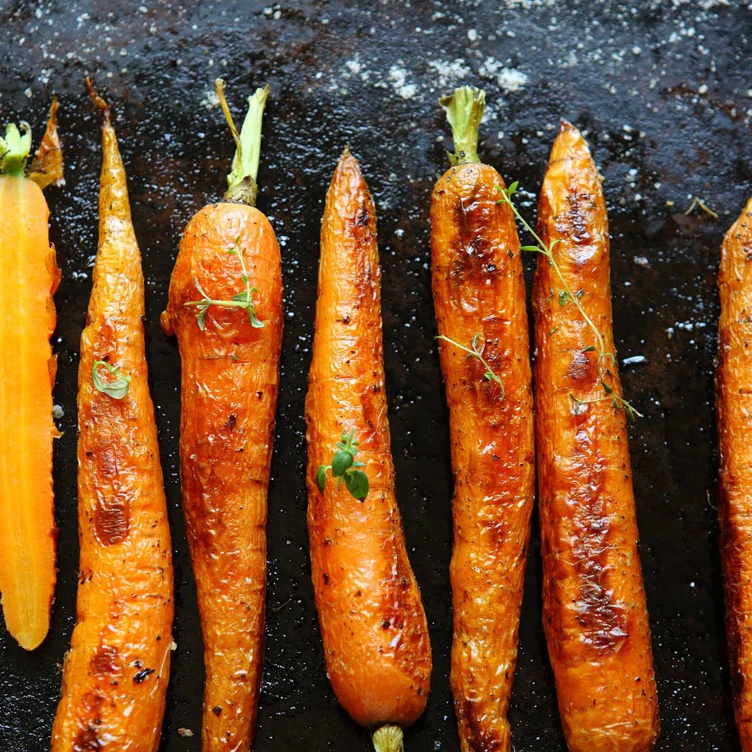 Row of glazed roasted carrots on a dark baking sheet, with sprigs of thyme for garnish.